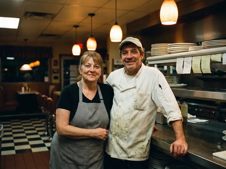 Two owners standing behind the pass of their diner